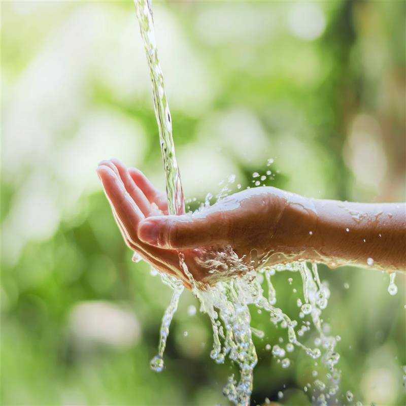 Clear water pouring over open hand against green sunlight background.