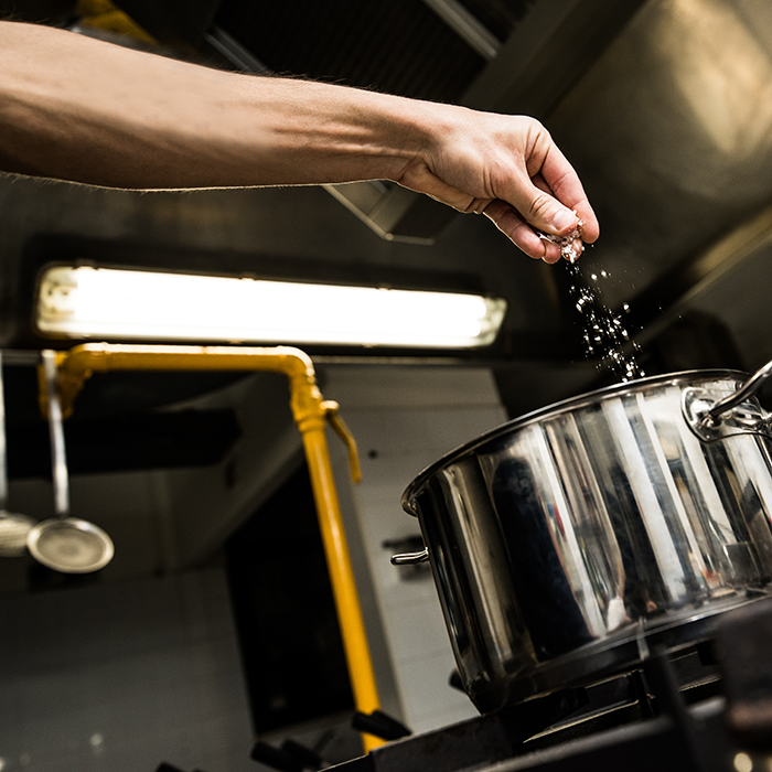 A hand adding salt to a pot on a stove burner in an industrial or commercial kitchen
