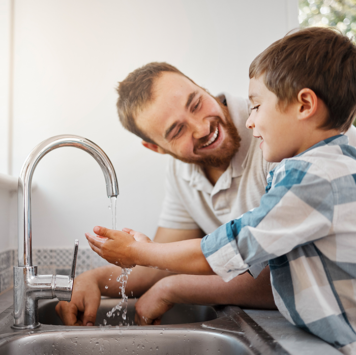 A man helping a young boy wash his hands at a kitchen sink under running water from a faucet.